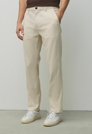 Man wearing beige straight-leg pants and white sneakers, standing with one hand in pocket against a plain background.