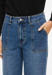 Blå høytlivs jeans i denim med knappelukking, to store fremlommer og kontrastsøm for ekstra detaljer og tekstur.