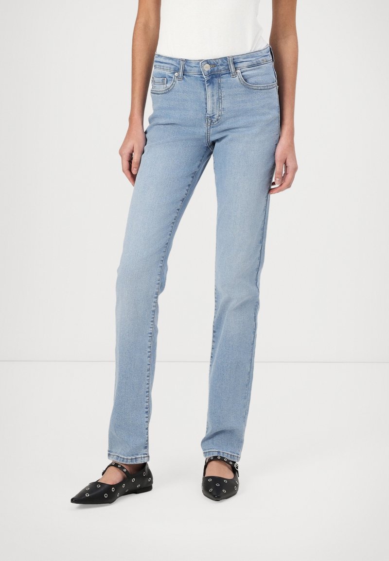 Femme portant un jean droit bleu clair et des ballerines noires à bouts pointus avec des détails œillets, debout sur un fond blanc.