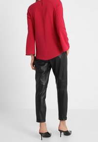 Rotes Langarmshirt mit Rundhalsausschnitt und Schlitz im Rücken, kombiniert mit schwarzen Lederanzughosen und schwarzen High Heels.