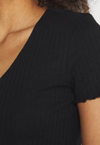 Top corto negro de manga corta con cuello en V, tejido texturizado y dobladillo en forma de onda en las mangas. Vista en primer plano de la prenda.