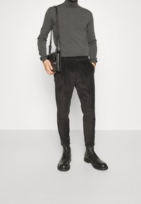 Mann trägt einen dunkelgrauen Rollkragenpullover, schwarze kordelartige Hose, schwarze Lederstiefel und trägt eine schwarze Lederumhängetasche.