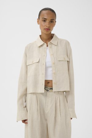 Femme portant une veste beige courte à boutons avec poches à rabat, pantalon large assorti, top blanc cropé et ceinture en peau de serpent.