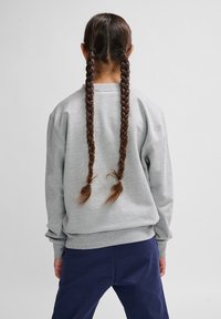 Felpa grigia con una vestibilità rilassata, collo rotondo e maniche lunghe. Presenta due lunghe trecce di capelli raccolte con elastici. Pantaloni navy sottostanti.