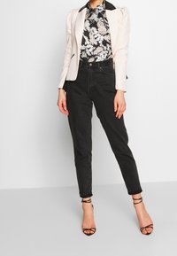 Blazer beige chiaro con colletto nero, blusa floreale e jeans neri a vita alta, abbinati a tacchi neri con cinturini.