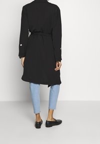 Czarny płaszcz trencz z paskiem, długimi rękawami i ozdobnymi guzikami, zestawiony z jasnoniebieskimi, skróconymi dżinsami oraz czarnymi mokasynami. Gładka tkanina i dopasowany krój.