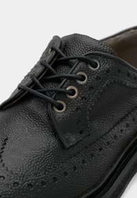 Chaussure en cuir noire à surface texturée, avec des détails brogues, six œillets et des lacets noirs. Bout rond et coutures contrastantes.