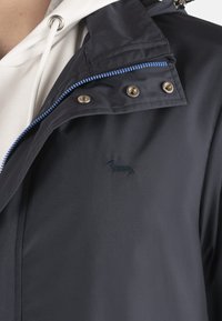 Marineblau wasserdichte Jacke mit Kapuze, ausgestattet mit blauem Reißverschluss, Metallbeschlägen und einem gestickten Dackel-Logo auf der Brust.