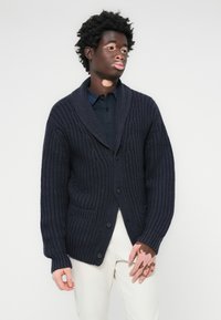 Mornarska pletena kardigan jakna s obodom, prednjim gumbima i dva džepa. Ima rebrastu teksturu i široke rukave.