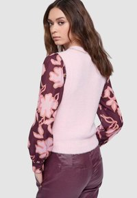 Maglione rosa senza maniche con una texture soffice, abbinato a maniche lunghe viola con motivo floreale. La modella indossa pantaloni aderenti color bordeaux.