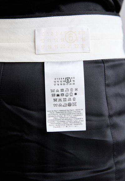 Melnā apģērba daļa ar baltu jostas nosaukuma etiķeti; ietver kopšanas instrukcijas un izmēru informāciju. Etiķetēs ir izdrukāti ikoni par veļas kopšanu.