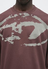 Bordo pulover s kapuco, na katerem je velik, abstrakten siv vzorec. Design z okroglim izrezom, mehka tekstura in vidni šivi na izrezu.