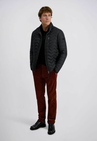 Een man in een zwarte gewatteerde jas, zwarte shirt, roestkleurige corduroypants en zwarte nette schoenen staat met zijn handen in zijn zakken.