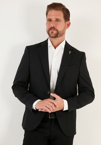 Musta bleiser, millel on kohandatud lõige, nervu krae, ühe nupuga kinnitamine ja taskurätt. Kantud valge särgi peal, millel on nähtavad nupud.
