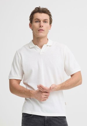 Hvid polo med korte ærmer, en klassisk krave og et tekstureret stof. Båret med mørke jeans, hænder placeret foran.