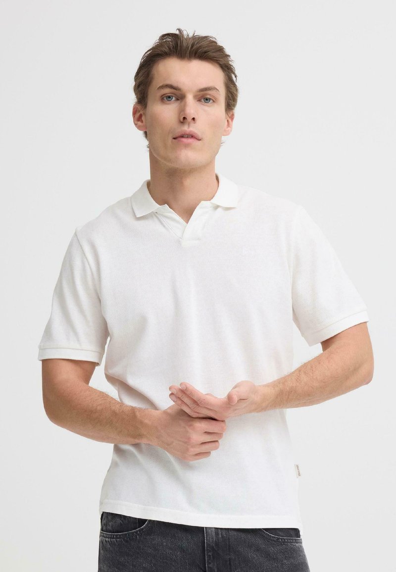 Polo blanc à manches courtes, col classique et tissu texturé. Porté avec un jean foncé, les mains placées devant.