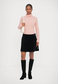 Lys rosa genser med høy hals, svart kordfløyelsfolderet skjørt og svarte knee-high støvler, kombinert med en liten hvit håndveske.