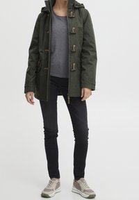 Osoba oblečená v olivově zeleném kabátu s kapucí, šedém tričku, černých skinny kalhotách a béžových teniskách stojící na jednobarevném pozadí.