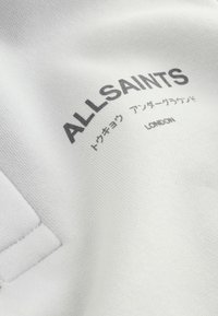 Bílá látka s nápisem "ALLSAINTS", japonskými znaky a nápisem "LONDON" vytištěnými černě na povrchu blízko okraje prošité kapsy.