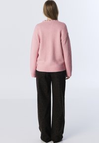 Różowy sweter robiony na drutach o miękkiej teksturze, z ribbowanym wykończeniem i ozdobnymi guzikami przy kołnierzu. Połączony z szerokimi spodniami w czarno-leopardzi wzór.