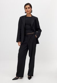 Schwarzer maßgeschneiderter Blazer kombiniert mit einem enganliegenden schwarzen Oberteil und lockeren schwarzen Hosen, ausgestattet mit eleganten, spitzen schwarzen Absätzen. Glatte Stoffqualität, minimalistisches Design.