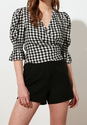 Vrouw draagt een zwart-wit geruit wikkelblouse met opgeblazen driekwartmouwen en zwarte shorts met hoge taille, staand tegen een effen lichte achtergrond.