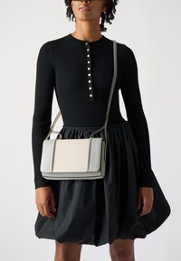 Borsa a spalla grigia e crema con design strutturato, texture liscia, bordi a contrasto e tracolla regolabile, indossata con un vestito nero a coste.