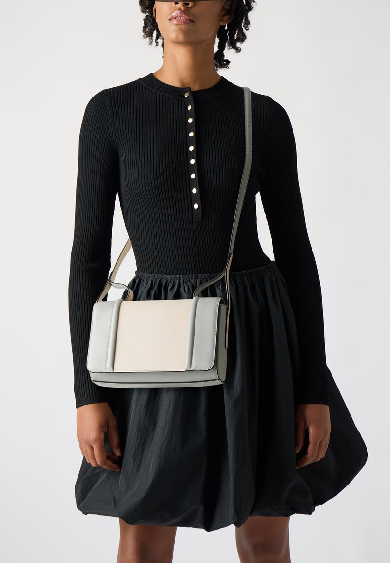 Borsa a spalla grigia e crema con design strutturato, texture liscia, bordi a contrasto e tracolla regolabile, indossata con un vestito nero a coste.