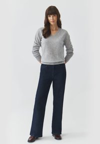 Grå V-hals sweater lavet af blødt stof; parret med mørkeblå, bredbenede denimjeans; model med hænderne i lommerne, stående på hvid baggrund.