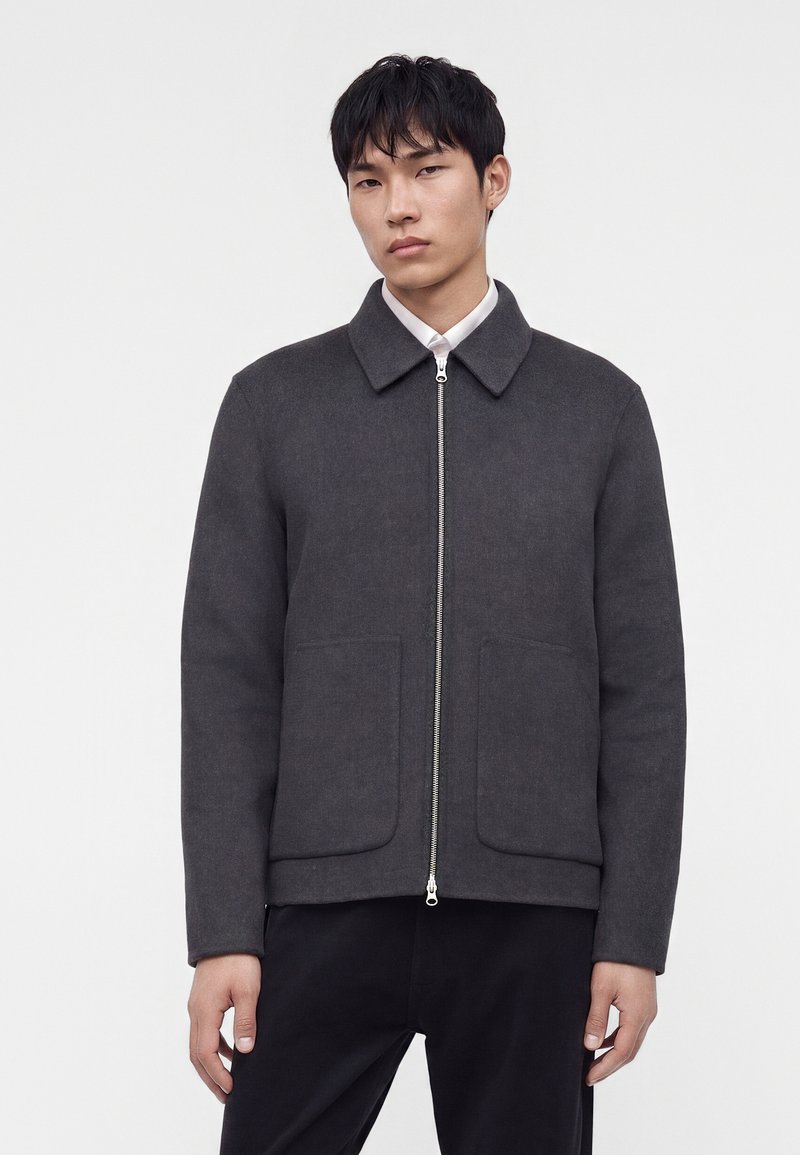 Jeune homme portant une veste zippée gris foncé avec de grandes poches devant et un pantalon noir, debout devant un fond clair uni.