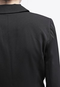 Veste noire sur mesure avec un tissu lisse, des épaules structurées et un design classique de revers. Le détail au dos montre des coutures nettes.