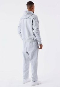 Sudadera gris clara con capucha y pantalones de chándal a juego, con puños acanalados y un logo sutil en la parte posterior, combinada con zapatillas blancas.