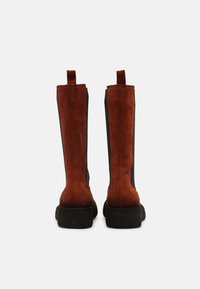 Brązowe zamszowe botki sięgające kostki z czarnymi elastycznymi panelami bocznymi, płaską gumową podeszwą i pętelkami z tyłu dla łatwego zakładania.