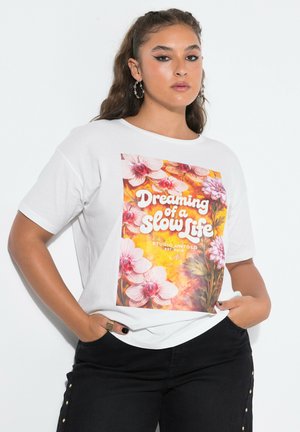 Jeune femme aux longs cheveux portant un t-shirt blanc avec un imprimé floral et le texte "Dreaming of a Slow Life,", associé à un pantalon noir.
