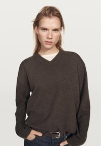 Žena s blond vlasy, která má na sobě tmavě hnědý svetřík s výstřihem do V přes bílý top, modré džíny a hnědý pásek, ruce v kapsách, neutrální pozadí.