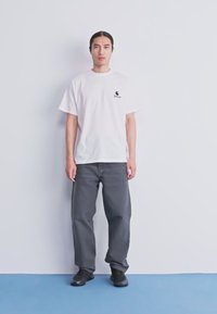 Hombre de pie contra una pared blanca, vistiendo una camiseta blanca con un pequeño logo negro, pantalones gris oscuro y zapatos negros sobre un suelo azul.