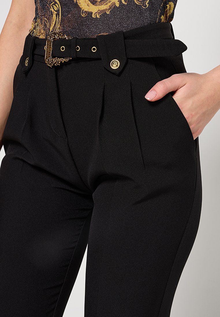 Pantalons noirs taille haute avec plis, dotés d'une large ceinture avec une boucle décorative en or et des boutons. Texture lisse et coupe ajustée.