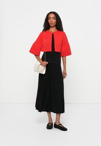 Giacca rossa corta con maniche a campana, abbinata a una gonna nera plissettata. Scarpe nere intrecciate e una borsa a tracolla color beige chiaro completano l'outfit.