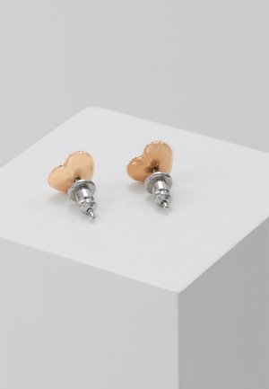 Petites boucles d'oreilles clous en forme de cœur doré avec des tiges en argent, présentées sur un socle blanc contre un fond gris.