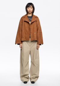 Bruine suedewanten met een oversized silhouet, drukknoopsluitingen en zijzakken, gecombineerd met losse beige broeken en zwarte enkellaarsjes.