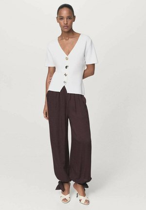 White short-sleeve V-neck top with five decorative buttons, paired with loose-fitting dark brown pants and cream open-toe heels.