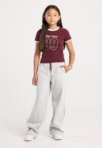 Ragazza giovane in piedi dritta, con una maglietta maroon "New York 89", pantaloni grigi chiari, sneakers bianche, con una mano in tasca e capelli lunghi.