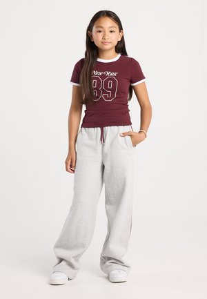 Jeune fille se tenant droite, portant un t-shirt bordeaux "New York 89", un pantalon de survêtement gris clair, des baskets blanches, avec une main dans la poche et de longs cheveux.