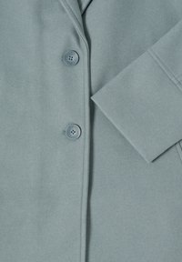 Ljusblå kappa med en slät textur, två knappar, slagkrage och en enkel manschett med ett rent avslut.