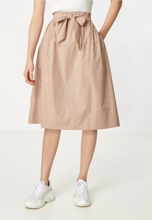 Femme portant une jupe midi beige et blanche à rayures verticales avec un noeud à la taille sur le devant et des baskets blanches, debout avec une main dans la poche.