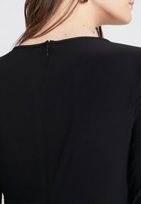 Musta mekko, jossa on sileä pinta, varustettuna piilotetulla vetoketjulla selkäpuolella ja yksinkertaisella pyöreällä kaula-aukolla.