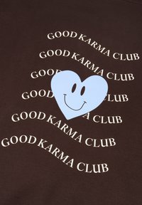 Tela marrón con un corazón azul claro sonriente, rodeado por el texto repetido "BUENA KARMA CLUB" en blanco.