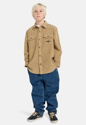 Light brown corduroy shirt with two chest pockets and black logo. Paired with loose blue denim jeans and black sneakers with white accents.