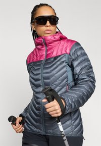 Eristetty takki, jossa on vaaleanpunainen yläosa ja tumma harmaa alaosa, jossa on korkea kaulus, vetoketjukiinnitys ja vino omplekuvio.