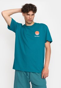 Teal katoenen T-shirt met een ronde halslijn en korte mouwen. De voorkant bevat een ronde grafiek in oranje en roze boven de tekst "Revolution."